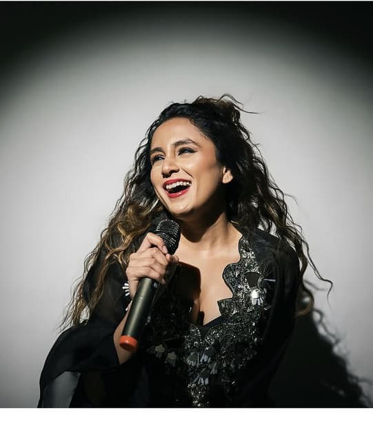 Kavya Limaye laughing mid-song, holding a microphone, in a black embellished outfit